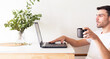 © Pablo - Retrato de un hombre adulto y apuesto caucásico sentado frente a su laptop y bebiendo una taza de café mientras la usa en un apartamento brillante y blanco con luz natural.