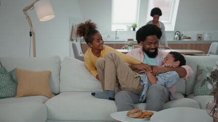 Wall Mural - Afro family in the living room. Father tickling his children, while mother is behind them in kitchen