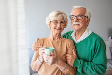 Enjoying time together. Side on waist up portrait of happy senior woman and man staying embracing near window at home interior. Portrait of senior couple standing near window