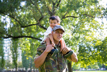 Loving Father Holding Son On Neck And Walking In City Park. Happy Caucasian Son Sitting On Neck Of Dad In Uniform, Hugging Him And Looking Away. Family Reunion, Fatherhood And Returning Home Concept