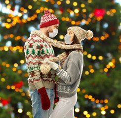 Wall Mural - winter holidays, pandemic and health concept - couple in sweaters wearing face protective medical masks for protection from virus disease over christmas tree lights on background