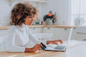 Wall Mural - Dark skinned lady with kinky hair in white typing on keyboard holding blue credit card enjoys shopping online in kitchen closeup