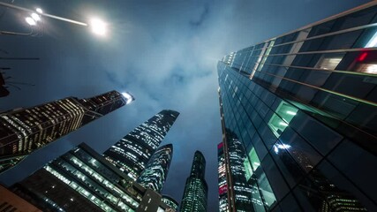 Wall Mural - Amazing motion timelapse of Moscow City Business Center on the night. View from the bottom to overhanging gigantic skyscrapers, twinkling with night lights, and the sky with fluffy clouds passing by.