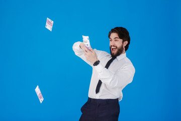 A young cheerful bearded businessman in a white shirt and black tie on a blue background splashes money. Crazy guy throws bills of money. Studio isolated portrait.