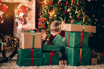 Canvas Print - Brother and sister are sitting with boxes in green packaging with gifts and a bow. Children in New Year's costumes waiting for a miracle. Against the background of a Christmas tree and fireplace