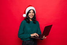 Attractive Young Woman With Santa's Christmas Hat Uses Laptop On Red Background.