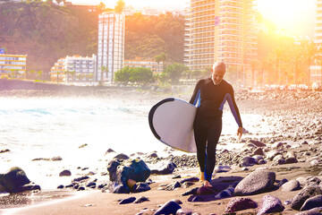 Active middle-aged man with surf board walking on the beach at sunset 