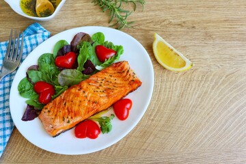 Grilled salmon with rosemary served with mixed fresh salad,heart shaped tomatoes,sliced lemon and olive oil with sliced rosemary in white bowl on white plate with wooden table background.Copy space