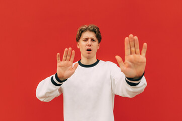 Wall Mural - Young man stands on a background of a red wall, looks at the camera with a surprised face and shows his palms gesture Stop. Guy shows his palms to the camera and says stop. Isolated.