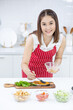 © narong - Asian beautiful woman with red apron smear oil and garnish on slicing fish on plate in kitchen with different types of bowl of ingredients on table. Concept of happiness of cooking in their house.