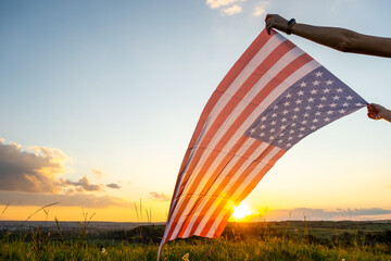 Human hands holding waving USA national flag in field at sunset.