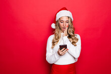 Surprised Young Woman In Santa Hat Using Mobile Phone Over Red Background