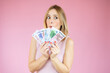 © Danko - Image of young funny woman standing over pink wall wearing casual clothes holding money. Looking aside.