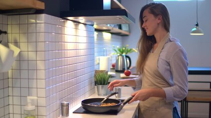 Wall Mural - Happy smiling attractive young cooking woman housewife preparing food in a frying pan on the stove for dinner at modern loft style kitchen