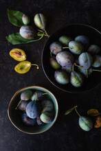 Overhead Shot Of Bowls Of Fresh Plums Against A Black Background.