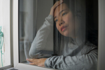 Wall Mural - young sad and depressed Asian Japanese girl scared and worried during covid19 lockdown and stay home quarantine looking through window feeling unhappy and stressed