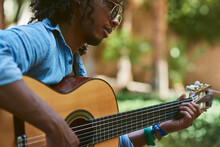 Young Musician With Classical Guitar Playing Under The Shade Of Some Trees In A Park.