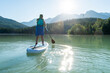 © Cavan Images - Back view of barefoot woman in dress and life vest riding SUP board on calm lake water on sunny summer day in mountains in British Columbia, Canada