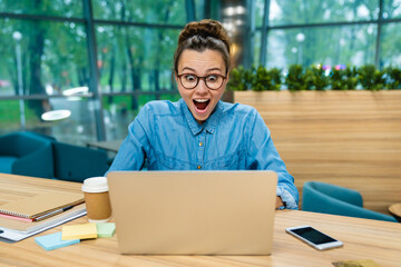 Surprised caucasian young girl wearing glasses using her laptop in modern office