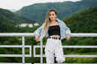© Cavan Images - Young woman with blonde braided hair wearing a denim jacket and white jean resting on a bridge