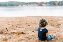 Little Toddler Girl With Curly Hair Sitting By A Lake With Pine Needle