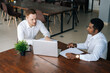 © dikushin - Two business partners discussing actively their startup project sitting at office desk, top view. African black businessman sitting ang talking his colleague while looking at laptop computer