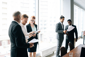 Wall Mural - Multicultural successful multi-ethnic coworkers reviewing documents background of large windows. Multiracial business team people and female corporate leader work together on paperwork