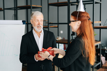 Business woman giving red gift box to smiling mature adult male leader during the Christmas, Happy New Year and Happy Birthday at office. Happy business lady congratulate to colleague.