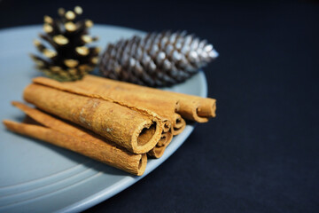 brown cinnamon sticks and two cones in gold and silver acrylic paint as Christmas decorations and on a gray plate on a black background side view . Christmas symbols