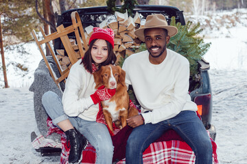 Wall Mural - Multiracial couple are sitting in the trunk of a car with a dog. A couple of lovers came to the forest for a Christmas tree.