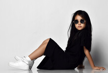 Full-growth Portrait Of Cool Asian Kid Girl In Stylish Black Dress, Round Sunglasses And White Sneakers Sitting On Floor Side To Camera Leaning Back On Her Hands. Trendy Children Fashion Concept