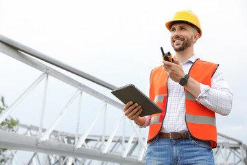 Professional engineer with tablet and walkie talkie near high voltage tower construction outdoors. Installation of electrical substation