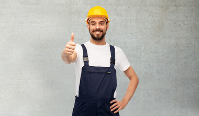 profession, construction and building concept - happy smiling male worker or builder in yellow helmet and overall showing thumbs up over grey concrete background