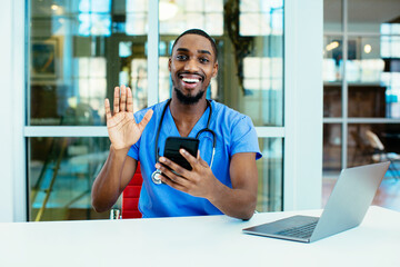 Wall Mural - Portrait of a smiling male doctor wearing blue scrubs uniform waving at camera, talk to patient online on phone