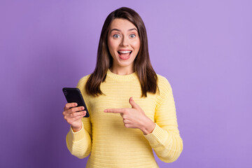 Poster - Photo portrait of excited woman holding phone in one hand pointing finger with open mouth isolated on vivid purple colored background