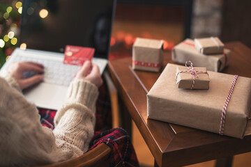 Wall Mural - Christmas. Woman in sweater holding credit card using laptop for making order sitting at table with packaging gift near fireplace and christmas tree. Online shopping concept