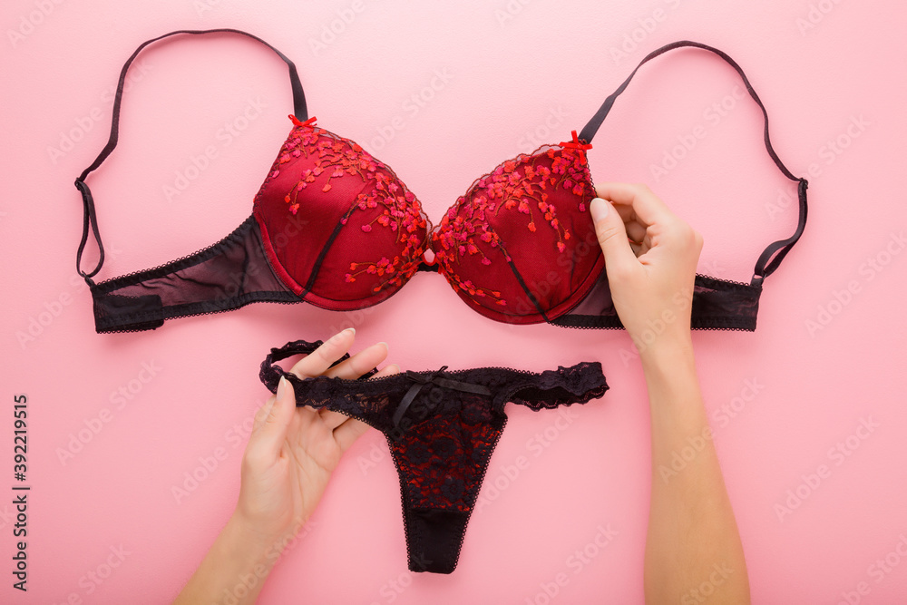 Young adult woman hands holding black red bra and string panties on light pink table background. Pastel color. Daily underwear. Closeup. Point of view shot. Top down view. - obrazy, fototapety, plakaty 