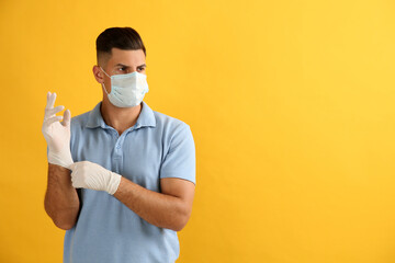 Poster - Man in protective face mask putting on medical gloves against yellow background. Space for text