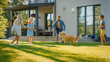 Smiling Beautiful Family Of Four Play Fetch Flying Disc With Happy Golden Retriever Dog On The Backyard Lawn. Idyllic Family Has Fun With Loyal Pedigree Dog Outdoors In Summer House Backyard