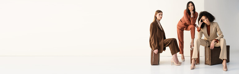 full length of multicultural women in trendy suits sitting on suitcases on white, banner