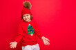 © Ivan Traimak - cool teenager in a red Christmas sweater fooling around against the background of a red wall, a warm hat and a sweater with a Christmas tree