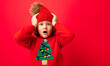 © Ivan Traimak - cool blond kid in warm hat and sweater with christmas tree on red background fooling around, christmas concept