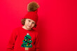 © Ivan Traimak - cool blond kid in warm hat and sweater with christmas tree on red background fooling around, christmas concept