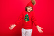© Ivan Traimak - cool teenager in a red Christmas sweater fooling around against the background of a red wall, a warm hat and a sweater with a Christmas tree