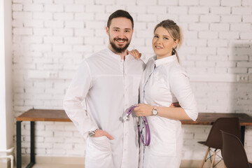 Poster - Portrait of smiling man and woman wearing white coats, looking at camera while standing in modern hospital room. on background white brick wall. Doctor standing upright while waiting for his team