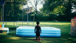 A young boy standing in a grassy backyard in the summer during quarantine using his imagination while playing in the sprinkler by the pool and trampoline