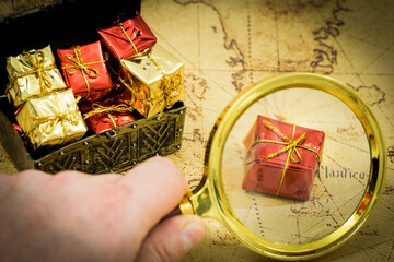 Aufkleber - the treasure hunt. A hand with a magnifying glass above a chest full of valuables and gifts, In the background an old map for adventure hunters