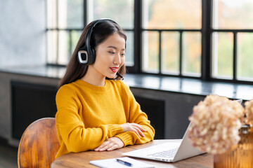 Asian woman in headset speaking by conference call and video chat on laptop in office, smiling chinese support service agent consulting customer. Female portrait looking at camera