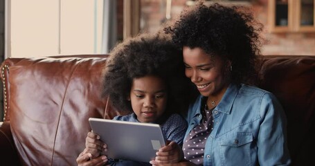 Wall Mural - Little girl and mother sit on couch with tablet device. African ethnicity family spend time on internet, enjoy educational web sites for kid development, modern tech usage for fun or education concept