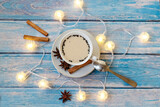 Fototapeta Dmuchawce - A Cup of cappuccino coffee, stars of anise and cinnamon sticks, a lighted garland on a blue wooden background. New year background
